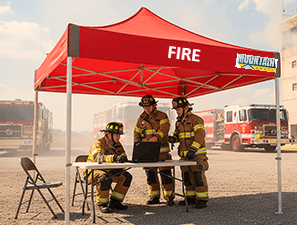 Three firefighters in turnout gear stand under a red canopy that says 'FIRE,' gathered around a table in a dusty lot with fire trucks in the background.