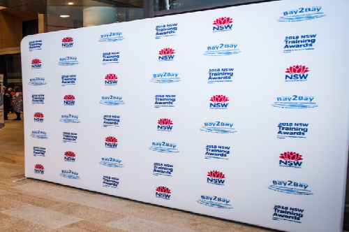 Indoor event backdrop featuring a white step-and-repeat banner with NSW logos and 'Bay2Bay' training awards repeated across.